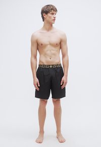 Zwarte zwemshorts met een gouden patroon op de tailleband, met een ontspannen pasvorm en een gladde textuur, gedragen door een mannelijke model die op blote voeten staat.