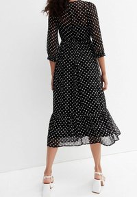 Schwarzes Kleid mit weißen Polka-Dots, 3/4-Ärmeln, gerüschtem Saum und tailliertem Schnitt. Durchscheinender, leichter Stoff mit durchgehenden weißen Punkten.