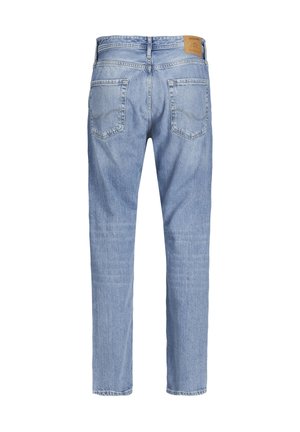 Homme portant un jean bleu clair, des bottes à lacets couleur beige et une chemise blanche, debout devant un fond clair uni.