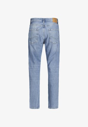 Mann mit hellblauen Jeans, hellbraunen Schnürstiefeln und weißem Hemd steht vor einem einfarbigen hellen Hintergrund.