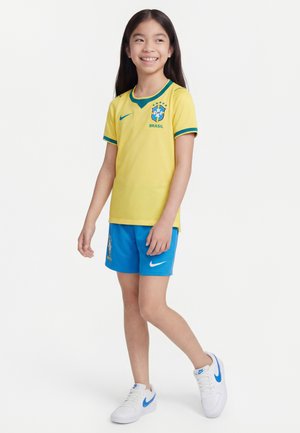 Fată zâmbitoare stând, purtând tricoul de fotbal al Braziliei galben și pantaloni scurți albaștri cu adidași albi Nike pe fundal alb.