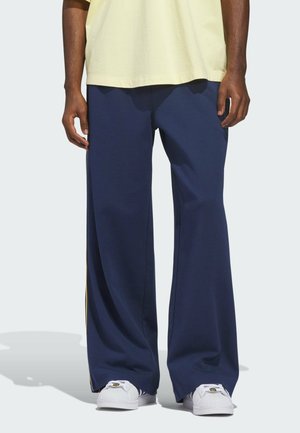 Marineblaue, weit geschnittene Hose aus weichem Stoff mit gelben Seitenstreifen, kombiniert mit weißen Sneakers und einem hellgelben T-Shirt.