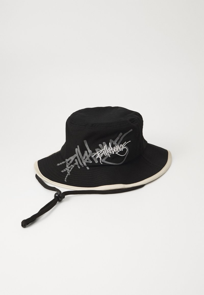 Black bucket hat made of cotton, featuring a wide brim with a cream accent, and graffiti-style logo in gray on the side. Includes a chin strap.