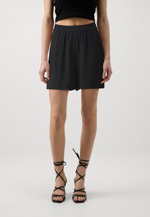 Zwarte shorts met elastische taille en een losser model, gecombineerd met zwarte sandaaltjes met banden die om de enkels zijn gewikkeld.