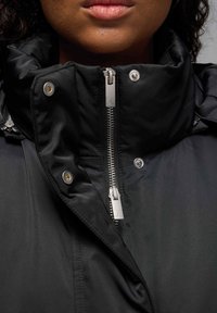 Giacca nera con colletto alto, zip metallizzata con logo, bottoni a scatto e texture liscia, che enfatizza un design elegante e moderno.