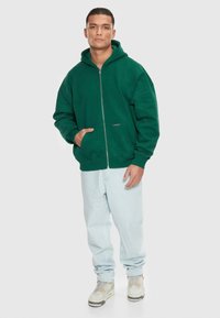 Grøn hættetrøje med frontlynlås, oversized pasform og en sidelomme, parret med lyseblå jeans og grå sneakers.