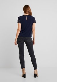 Navy top with lace short sleeves, featuring a back keyhole detail. Paired with dark grey skinny jeans and beige block-heeled shoes.