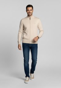 Suéter beige claro con cremallera en un diseño ajustado, combinado con jeans azul oscuro y zapatillas beige. Textura suave, mínima presencia de marca visible.