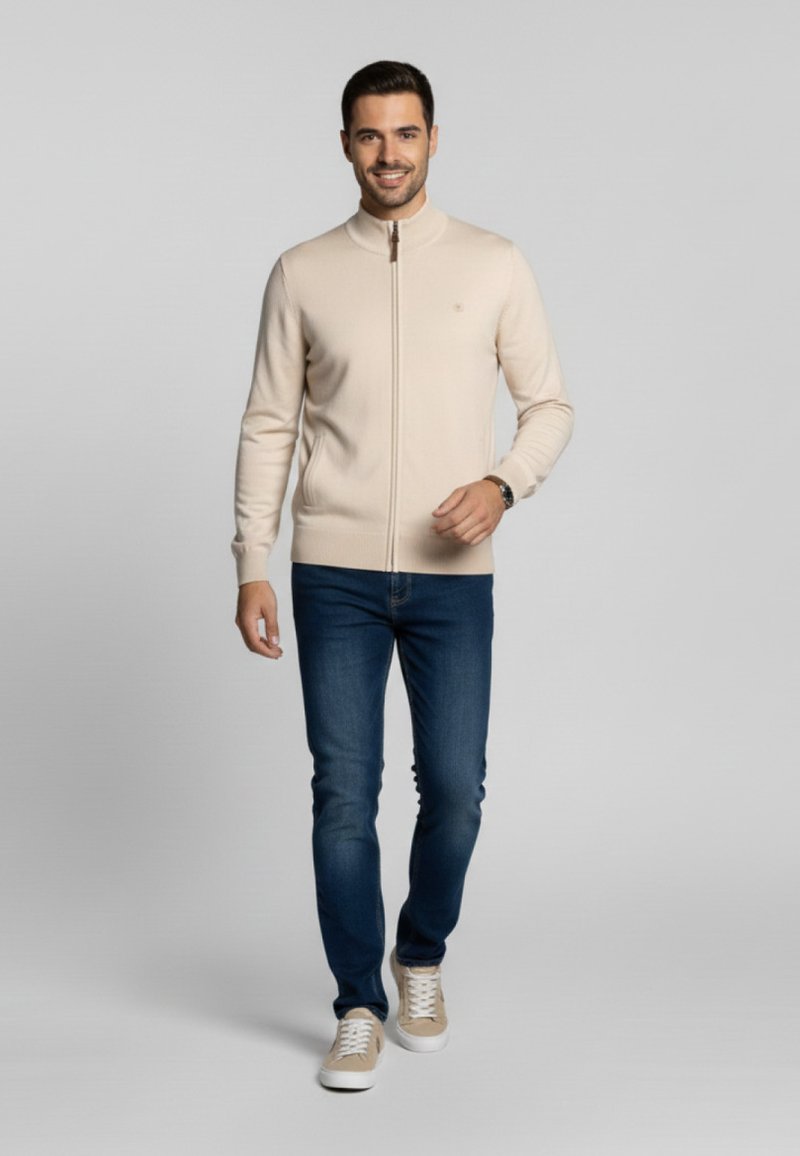 Suéter beige claro con cremallera en un diseño ajustado, combinado con jeans azul oscuro y zapatillas beige. Textura suave, mínima presencia de marca visible.