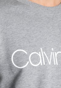 Felpa grigia realizzata in tessuto misto cotone con un grande logo bianco "Calvin". Il collo a costine aggiunge texture.