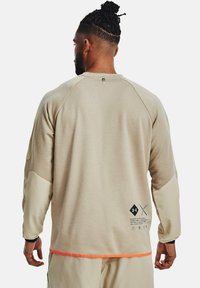Uomo con i capelli intrecciati che indossa una camicia beige a maniche lunghe con toppe color sabbia sui gomiti e bordi arancioni, visto da dietro.