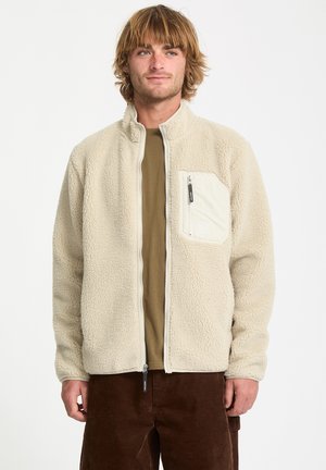 Cream-colored fleece jacket with a high collar, zippered front, and chest pocket. Soft texture and relaxed fit, worn over an olive shirt.