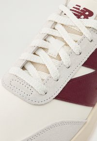 Fehér sneaker, bordó bőr díszítéssel, texturált velúr felsőrésszel, hálós elemekkel és sima krémszínű fűzőkkel. Kiszolgált varrási részletek láthatók.