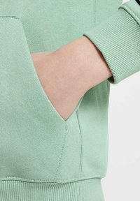 Ljusgrön sweatshirt med ribbad mudd och känguruficka. Tyget har en mjuk struktur med subtila stickade detaljer.