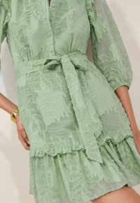 Leicht grünes Kleid mit bestickten Blättermustern, Knopfleiste, tailliertem Bund mit Bindeband und gerüschtem Saum. Texturiertes, durchscheinendes Gewebe.