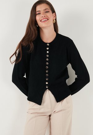Black knitted cardigan with a round neckline, silver buttons down the front, and a slightly curved hem. Worn over light-colored trousers.