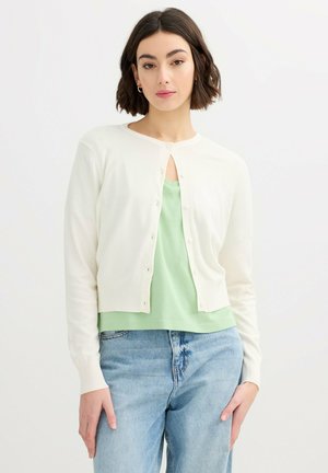 Mujer con el pelo corto y oscuro, que lleva un cárdigan blanco abotonado sobre una camiseta verde clara y vaqueros azul claro, de pie frente a un fondo blanco liso.