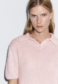 Světlá růžová pletená polokošile s jemnou texturou, krátkými rukávy a klasickým límcem s knoflíkovou légou a mírnou průhledností.