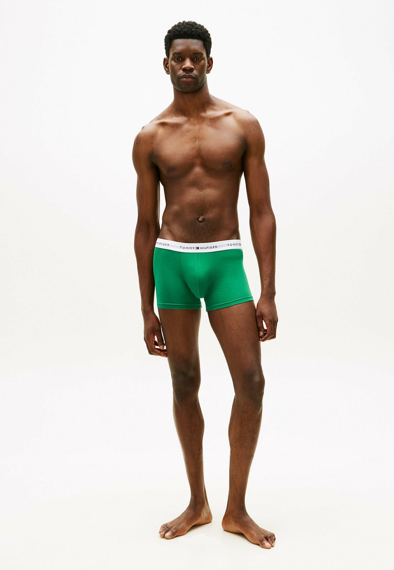 Giovane uomo in piedi a piedi nudi davanti a uno sfondo semplice, indossa boxer verdi Tommy Hilfiger con elastico bianco in vita.
