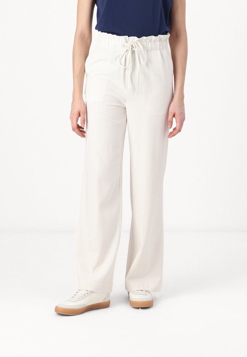 Pantaloni bianchi a gamba larga con vita elastica e cordoncino regolabile, realizzati in un materiale leggero. Abbinati a scarpe da ginnastica beige.