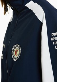 Navy blue windbreaker with white accents, full zip, stand collar, and embroidered patch detailing. Smooth texture and lightweight material.