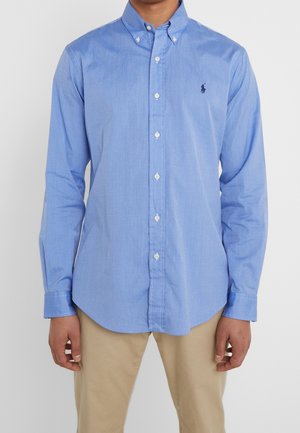 Muž v světle modré košili s límečkem a béžových kalhotách stojí před bílým pozadím.