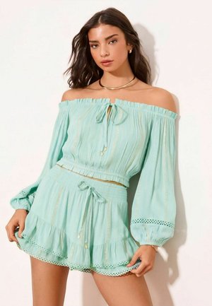 Junge Frau trägt ein hellblaues Off-Shoulder-Top mit langen Ärmeln und passende hoch taillierte Shorts mit Rüsch- und Spitzendetails.