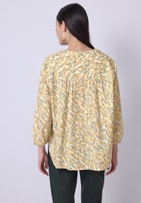 Blouse florale jaune avec des manches longues bouffantes, en tissu doux, et un détail froncé au dos. Un pantalon vert complète la tenue.