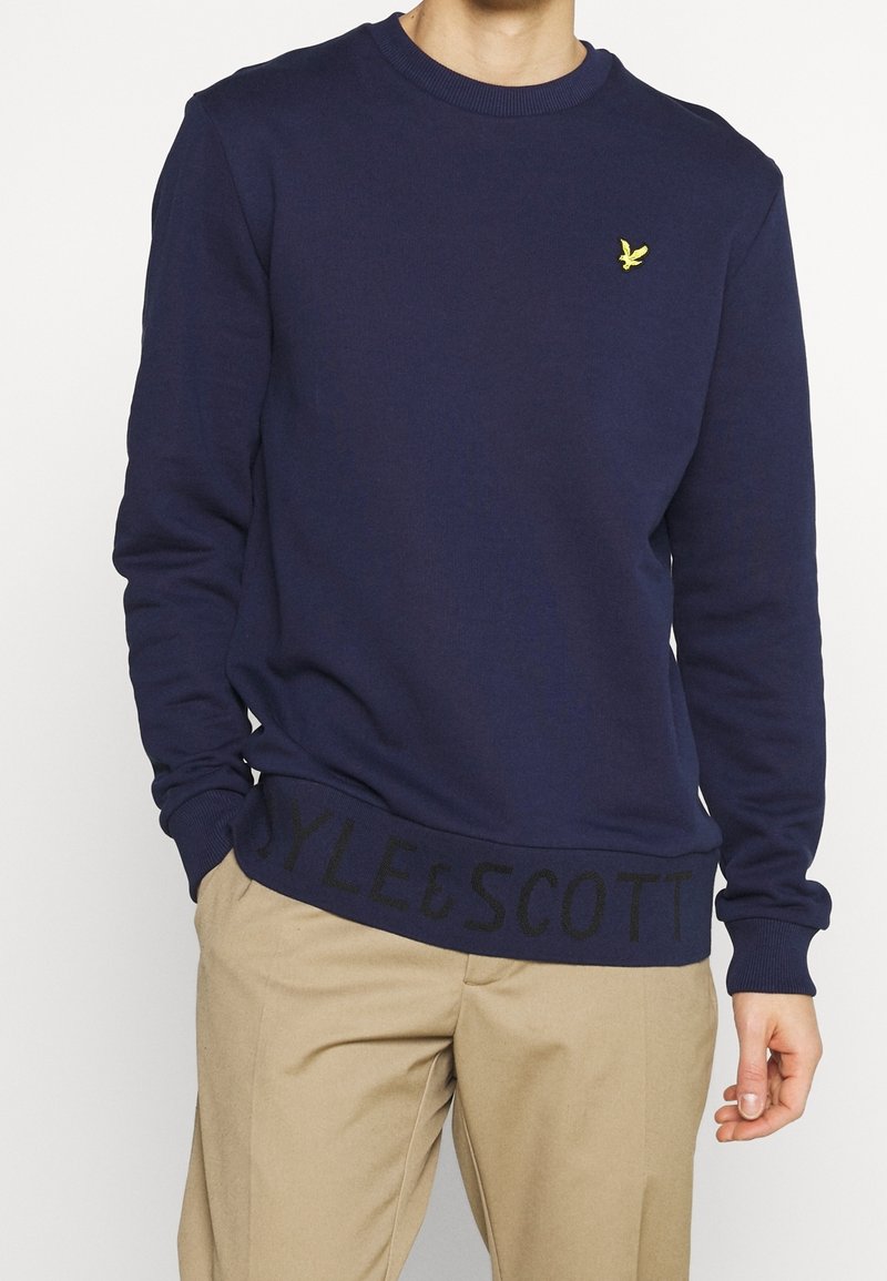 Sudadera de cuello redondo color azul marino en una tela suave, con puños y dobladillo de canalé. Presenta un pequeño logo amarillo y una banda negra con el texto "STYLE&SCOTT".