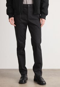 Pantalones negros de tejido suave y mate con un diseño de pierna recta, que presentan un cierre delantero con botón y bolsillos laterales. Llevados con zapatos negros.