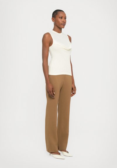 White sleeveless top with draped neckline, paired with brown straight-leg trousers and white pointed-toe shoes. Smooth textures, minimalist design.