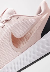 Světle růžová sportovní bota s texturovaným síťovaným svrškem, výrazným logem Nike v metalickém provedení a bílou gumovou podrážkou se vlnkovým vzorem.