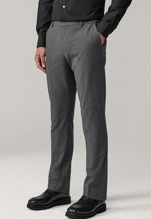 Szürke, oldalzsebes hivatalos nadrágot és fekete bőr elegáns cipőt viselő férfi, egyszínű háttér előtt áll.