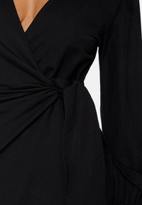 Zwarte wikkeljurk met lange mouwen, een diepe V-hals en een verzamelde taille met een knoopdetail. De stof heeft een soepele, matte textuur.