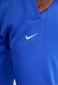 Blue athletic pants made of a smooth, lightweight fabric featuring a white Nike logo on the left thigh and stitched detailing along the seams.