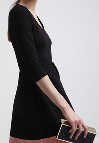 Robe noire à manches longues avec un corsage ajusté et une jupe évasée. Le mannequin tient une pochette scintillante de couleur sombre avec un cadre doré, posée sur un fond neutre.