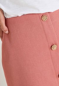 Jupe rose à côtes avec une finition texturée, dotée d'accents de boutons tressés en or et d'une couture latérale. Des mains visibles tenant la taille.