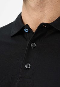 Zwarte polo met een textuurstof, een spitse kraag met één blauwe knop en drie zwarte knoppen, en een strak ontwerp.