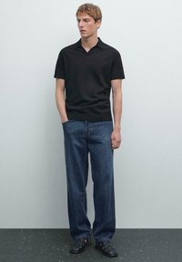 Mladý muž stojící oblečený v černém polo tričku s krátkým rukávem, modrých rovnějších džínách a černých sandálech se zakrytými prsty, před jednobarevnou zdí.