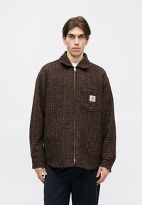 Brun og orange ternet zip-up jakke med krave, der har en frontlomme og et logo patch. Skåret løst for et afslappet fit.