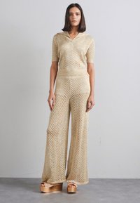 Combinaison tricotée rayée dorée et crème avec des manches courtes et un pantalon large, présentant un col en V. Assortie avec des chaussures compensées chunky.