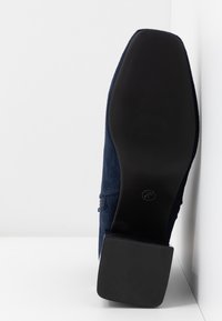 Chaussure en daim marine à semelle noire, debout contre un mur blanc, affichant la taille 41/37 marquée sur la semelle.