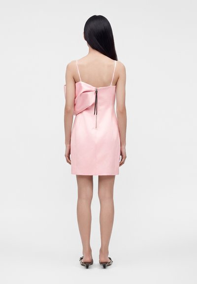 Femme aux longs cheveux noirs portant une robe rose sans manches avec un grand nœud et une fermeture éclair visible dans le dos, debout devant un fond uni.