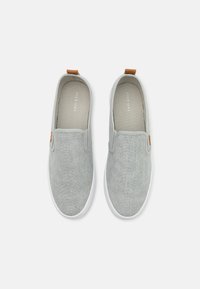 Világosszürke slip-on cipők, textúrált szövetfelsővel, fehér gumitalppal és egy kis barna bőr díszítéssel a sarkon.