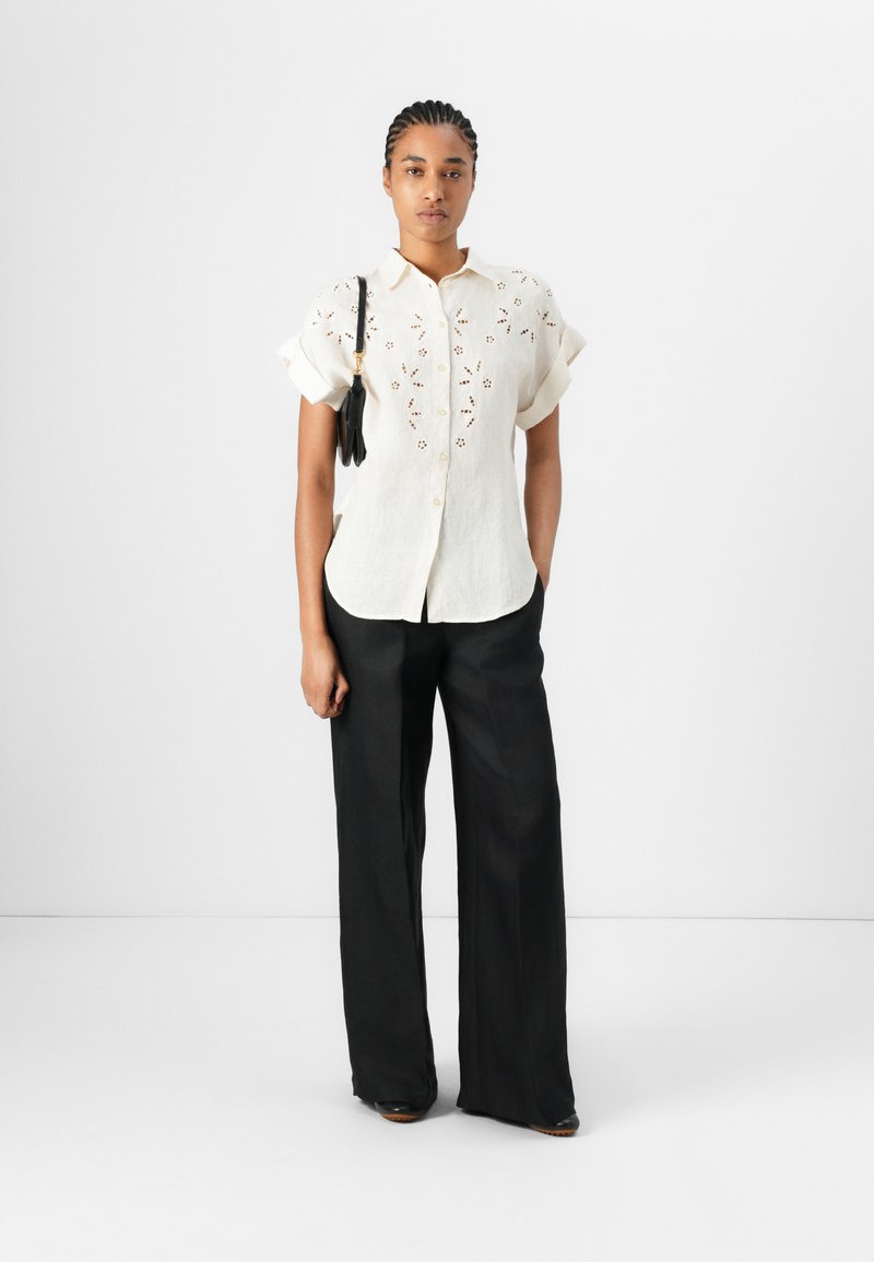 Helles Elfenbeinhemd mit floralen Stickereien, kurzen Rüschensleeves und einer Knopfleiste, kombiniert mit weiten schwarzen Hosen.