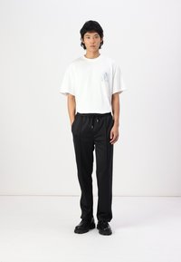 Tricou alb cu mânecă scurtă și design imprimat; pantaloni negri cu șnur, material neted, formă conică; model în picioare pe un fundal deschis.