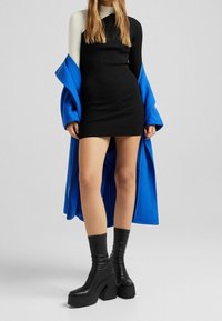 Robe mini noire à côtes avec manches longues ; portée sous un manteau bleu vif. Le mannequin porte des bottes à talons larges noires, mettant en valeur un design élégant.