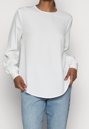 Weißes Langarmblusenshirt aus glattem Stoff, mit Rundhalsausschnitt, elastischen Bündchen und abgerundetem Saum. Kombiniert mit hellblauen Jeans.