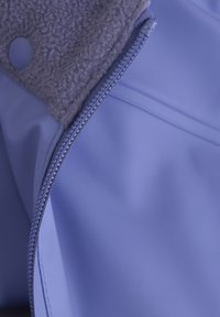 Ljuslila kappa med en mjuk grå fleecekrage. Har en metallisk dragkedja och textiler som framhäver både släta och fluffiga element.