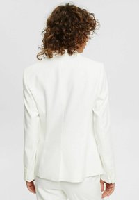 Kobieta z kręconymi włosami widziana z tyłu, ubrana w białą skrojoną marynarkę i pasujące białe spodnie na jednolitym tle.